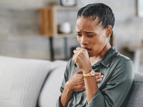 Woman sitting on sofa at home, frowning and pressing on chest, coughing into palm